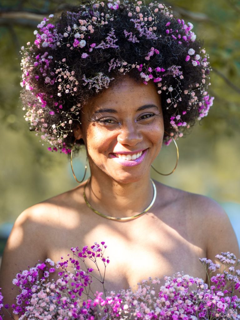 Erblüht Shooting Body and Soul Stories Frau mit fröhlichem Lächeln, Blumen-Afro und Blumenkranz vor natürlichem Hintergrund.