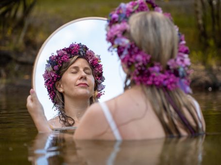 Erblüht Shooting Selbstliebe Coach Frau mit Blumenkranz, sitzt im Wasser und blickt in einen Spiegel.