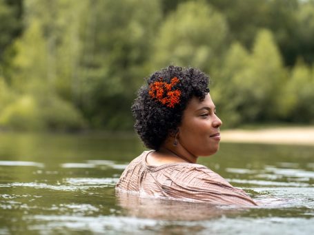 Erblüht – natürliche Frauenporträts in der Natur Frau mit Afro-Haaren und roten Blumen im Haar steht im Wasser, umgeben von Bäumen – natürliches Frauenporträt